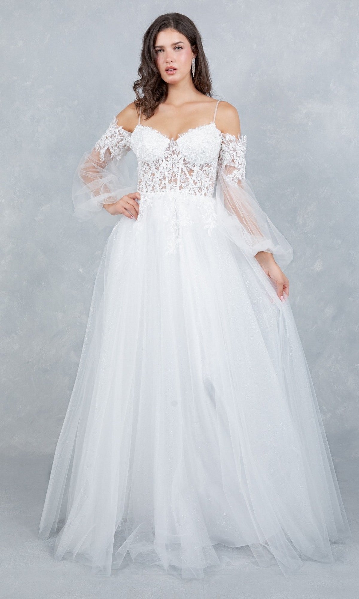 A woman stands against a light gray textured background wearing the Cinderella Couture 4234 long formal dress, featuring a white, off-the-shoulder design with a lace applique corset bodice and sheer tulle sleeves.